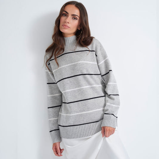 A woman wearing a light gray knit mock-neck sweater with horizontal black and white stripes, paired with a white skirt, standing against a plain white studio background.