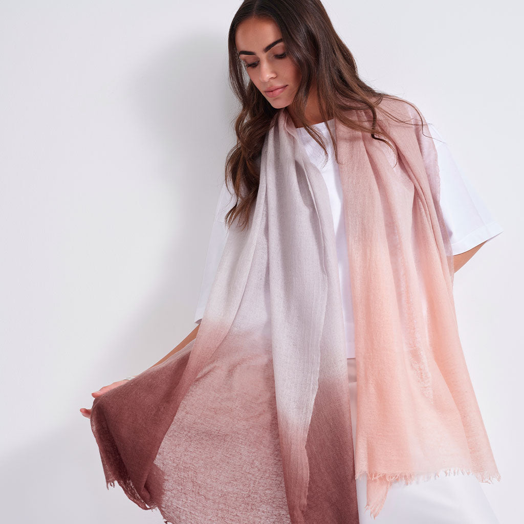 A woman wearing a lightweight, semi-sheer scarf with an ombré color transition from white at the top to a dusty rose pink at the bottom, styled over a white t-shirt against a plain white background.
