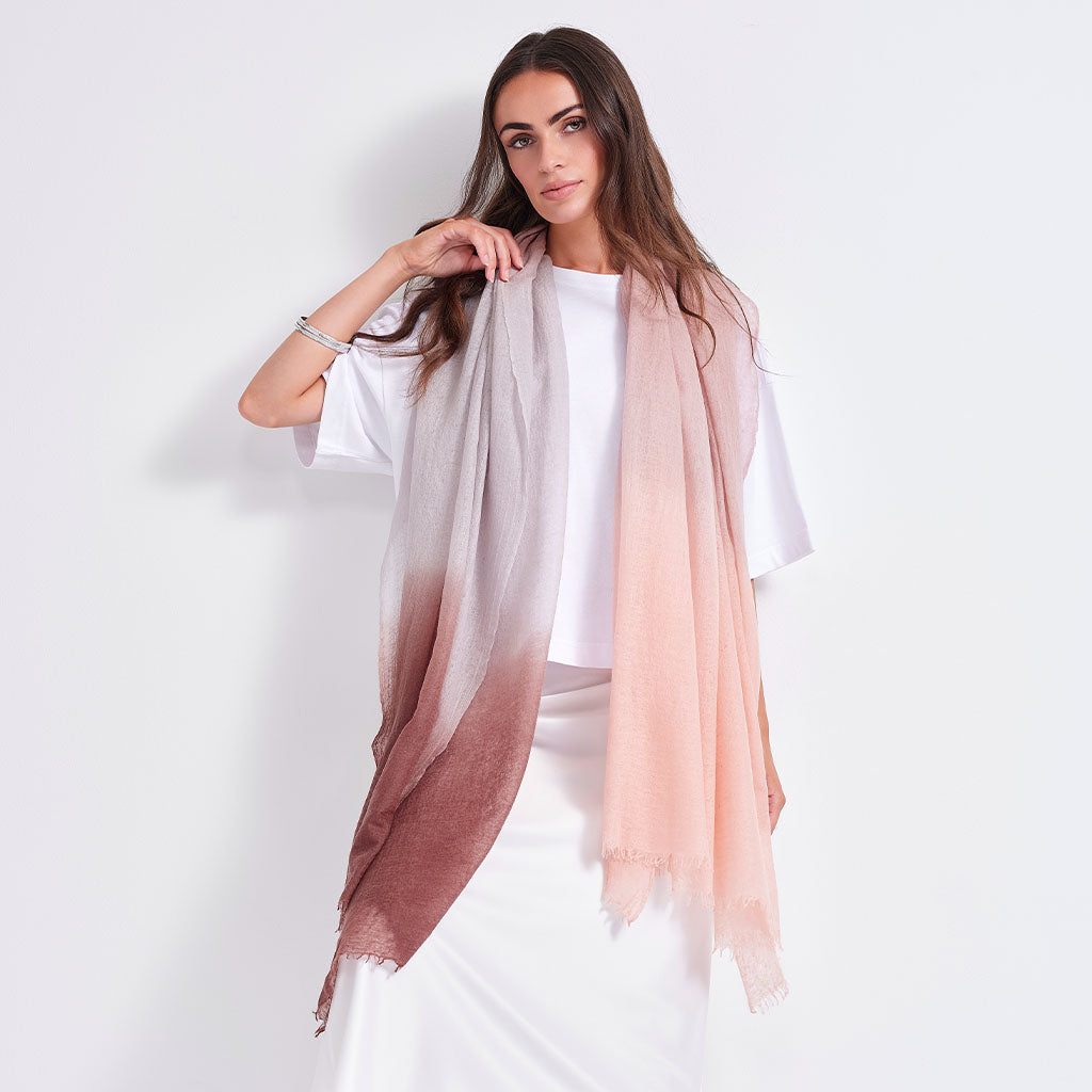 A woman modeling a lightweight gradient scarf that transitions from dusty rose to soft blush pink, worn over a white top and skirt against a plain white studio background.