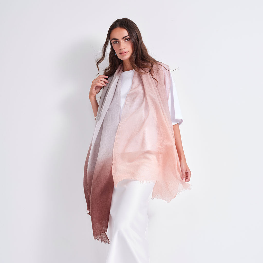 A woman models a lightweight, semi-sheer scarf with a vertical ombré transition from white at the top, through dusty pink in the middle, to a muted rose-brown at the bottom, worn over a white top and white trousers against a plain white studio background.