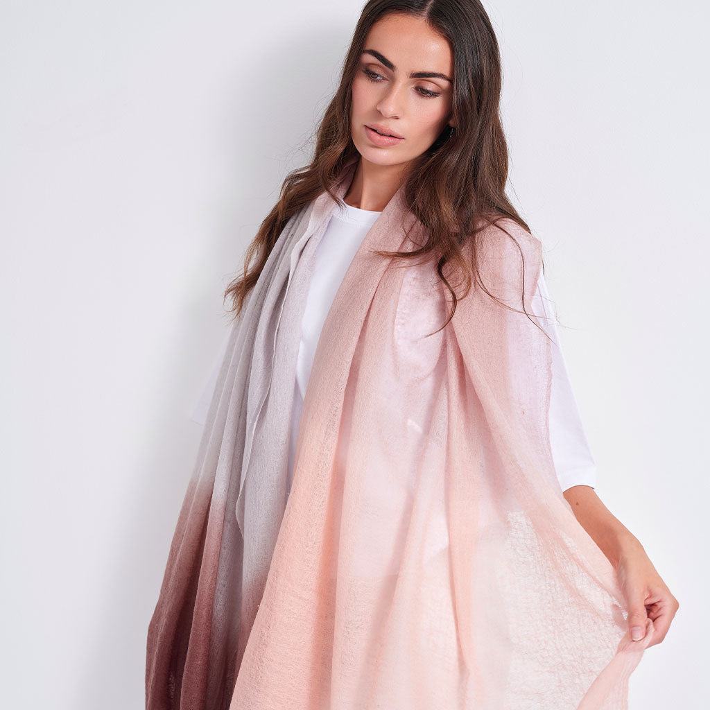 A woman modeling a lightweight, semi-sheer pink ombre drape or shawl over a white t-shirt, set against a plain light gray studio background.