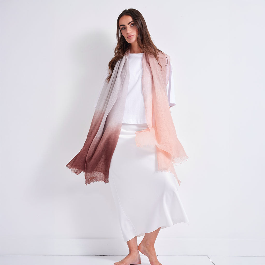 A woman stands barefoot against a plain white studio background, wearing a white top and white skirt or dress, with a sheer draped scarf or shawl in a soft gradient from pale blush pink at the top to dusty rose and mauve-brown at the bottom.