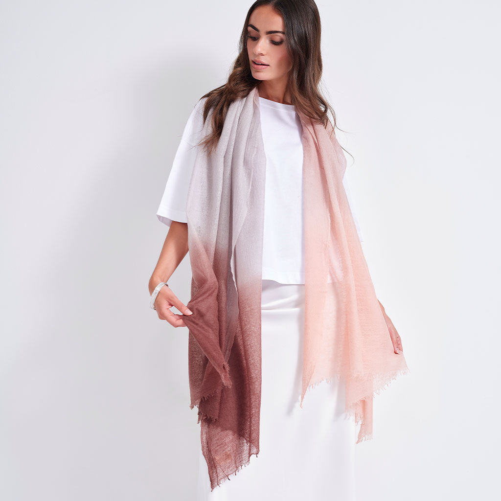 A woman models a lightweight, semi-sheer scarf with a vertical ombre gradient transitioning from light gray at the top to dusty rose at the bottom, styled over a white top and white skirt against a plain light gray background.