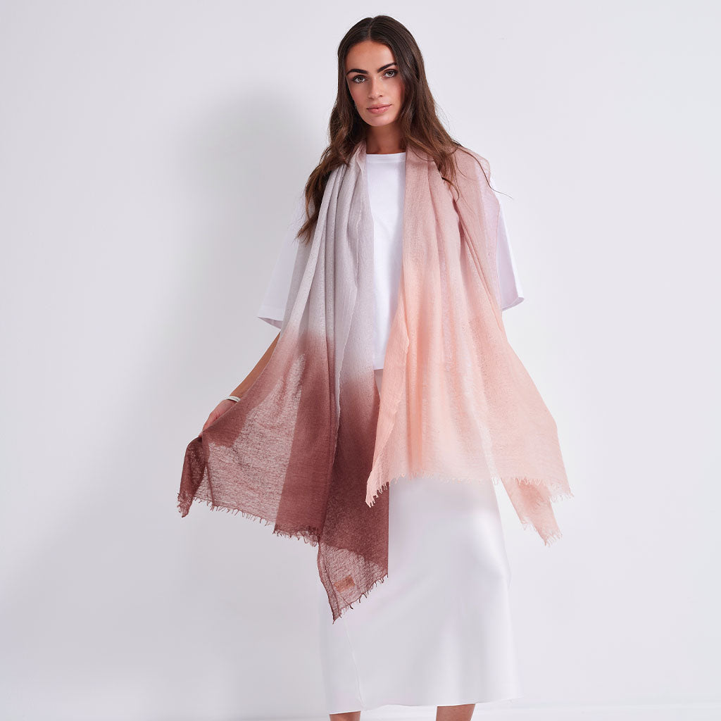 A woman modeling a lightweight gradient scarf that fades from white at the top to a soft peach-pink and dusty rose at the bottom, styled over a white dress against a plain white studio background.