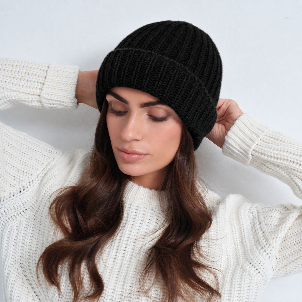A woman wearing a black ribbed knit beanie and a beige knit sweater, posing against a light gray studio background.
