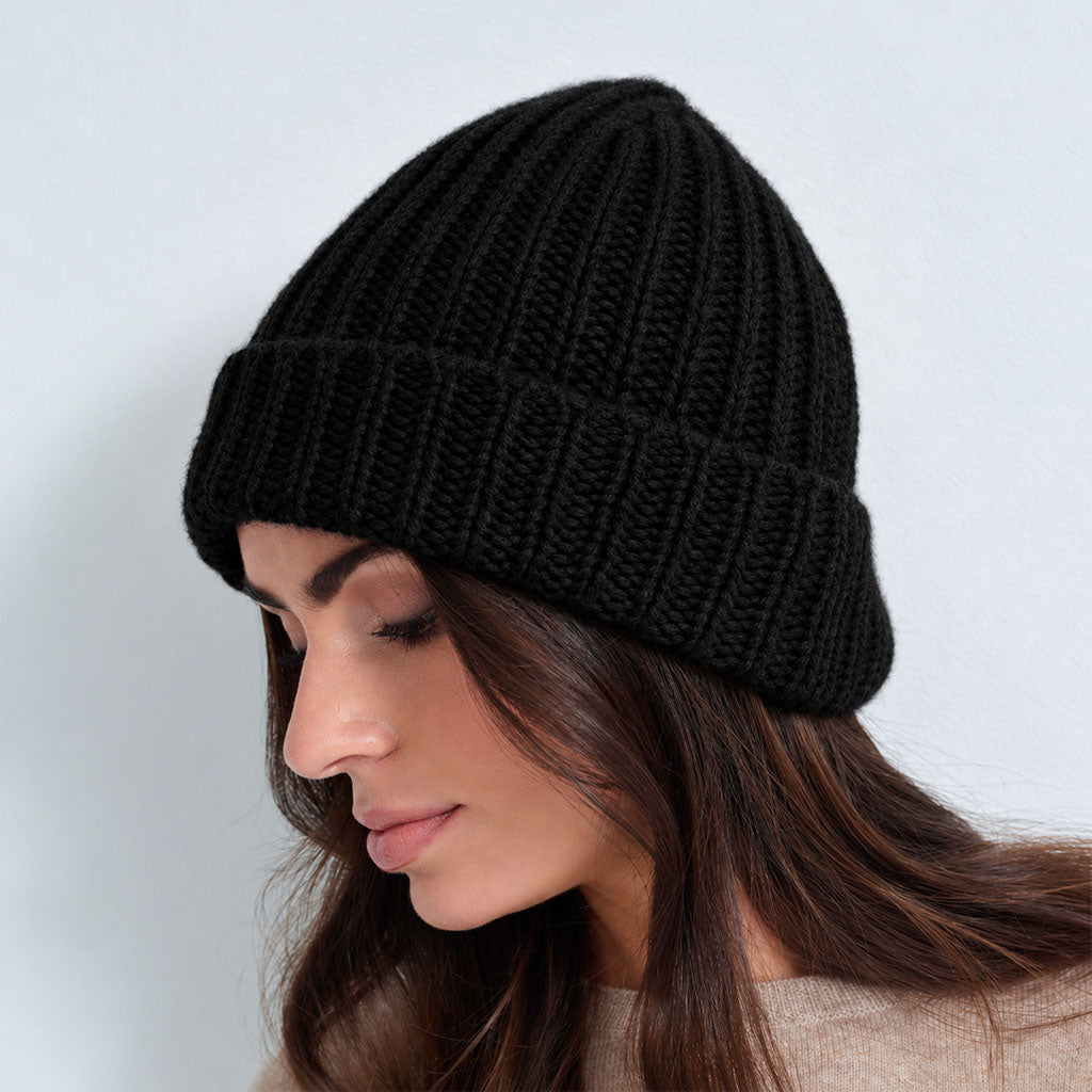 A woman wearing a black ribbed knit beanie and a beige knit sweater, posing against a light gray studio background.