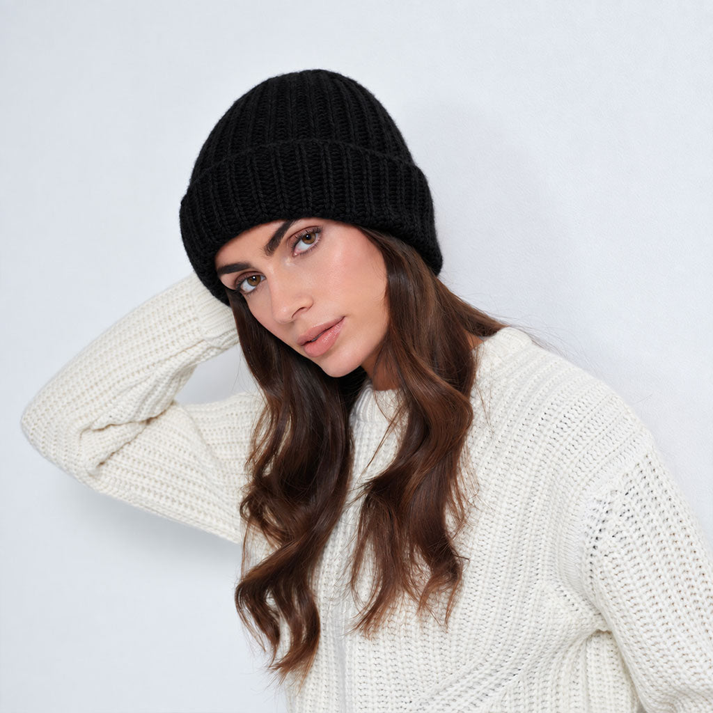 A woman wearing a black ribbed knit beanie and a beige knit sweater, posing against a light gray studio background.