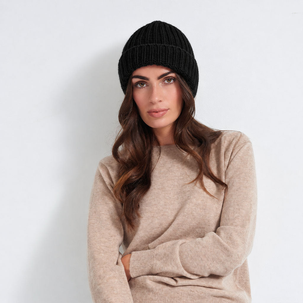 A woman wearing a black ribbed knit beanie and a beige knit sweater, posing against a light gray studio background.
