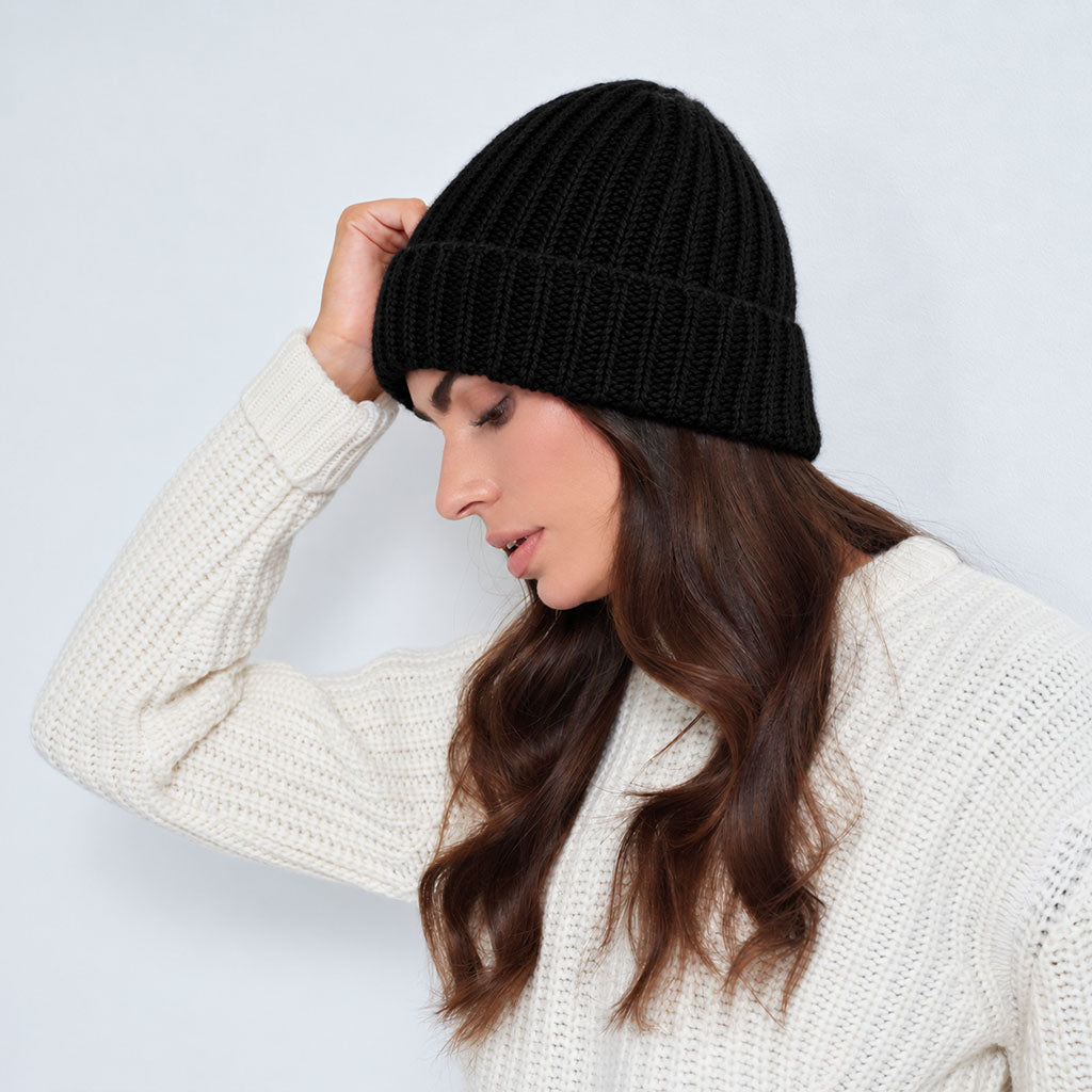 A woman wearing a black ribbed knit beanie and a cream knit sweater, posing against a light gray studio background.