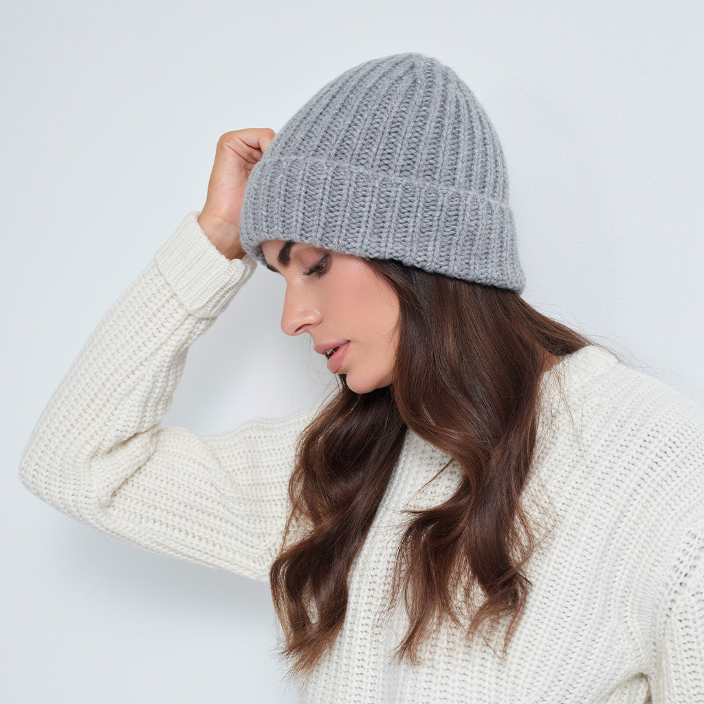 A woman wearing a light gray ribbed knit beanie and a cream-colored knit sweater, posing against a plain light gray background.
