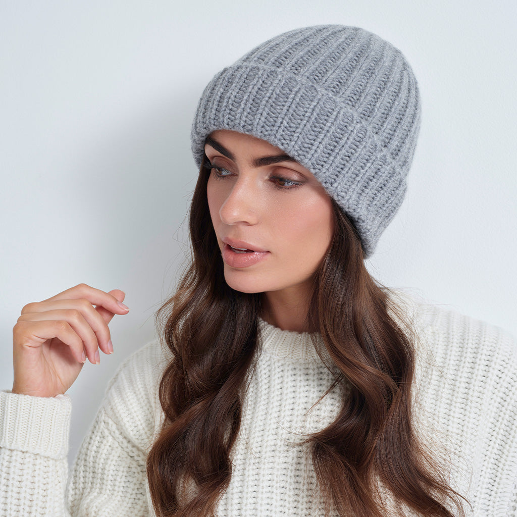 A woman wearing a light gray ribbed knit beanie and a cream-colored knit sweater, shown from the chest up against a plain light gray background.