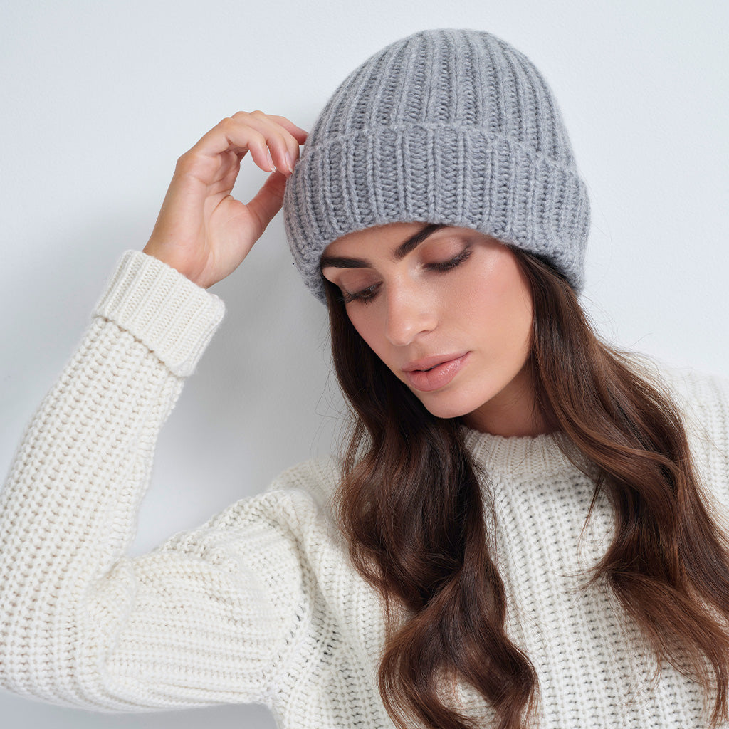 A woman wearing a light gray ribbed knit beanie and a cream-colored waffle-knit sweater, posing against a light gray background.