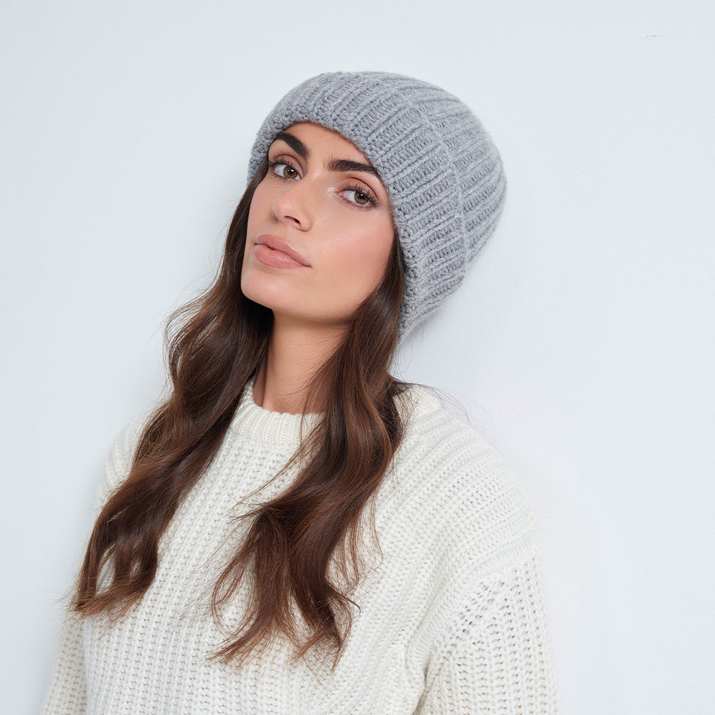 A woman wearing a light gray ribbed knit beanie and a cream-colored knit sweater, posing against a plain light gray background.