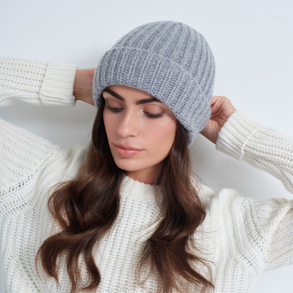 A woman wearing a light gray ribbed knit beanie and a cream-colored knit sweater, posing against a plain light gray background.
