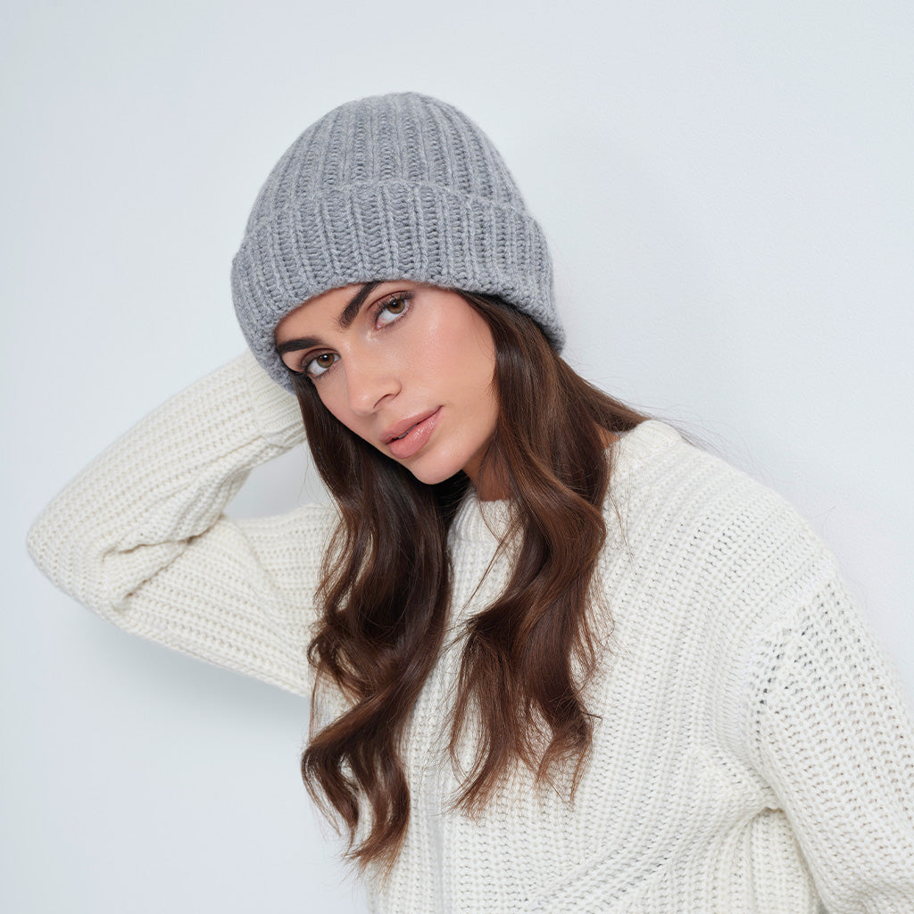 A woman wearing a light gray ribbed knit beanie and a cream-colored knit sweater, posing against a plain light gray background.