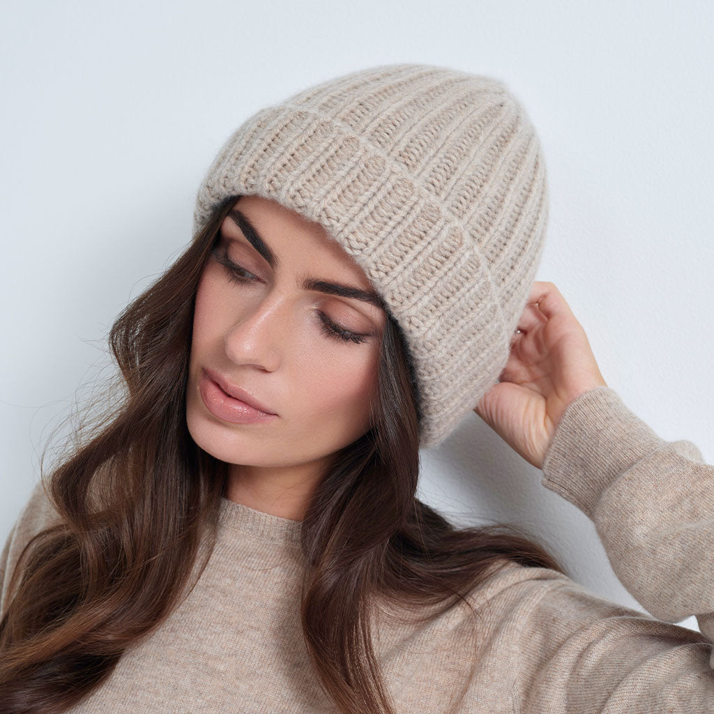 A woman wearing a beige ribbed knit beanie and a matching beige sweater, posing against a light gray background.