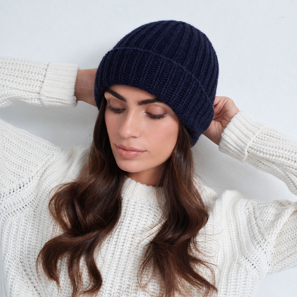 A woman wearing a dark navy blue ribbed knit beanie and a white textured knit sweater, posing with her hands behind her head against a plain white background.