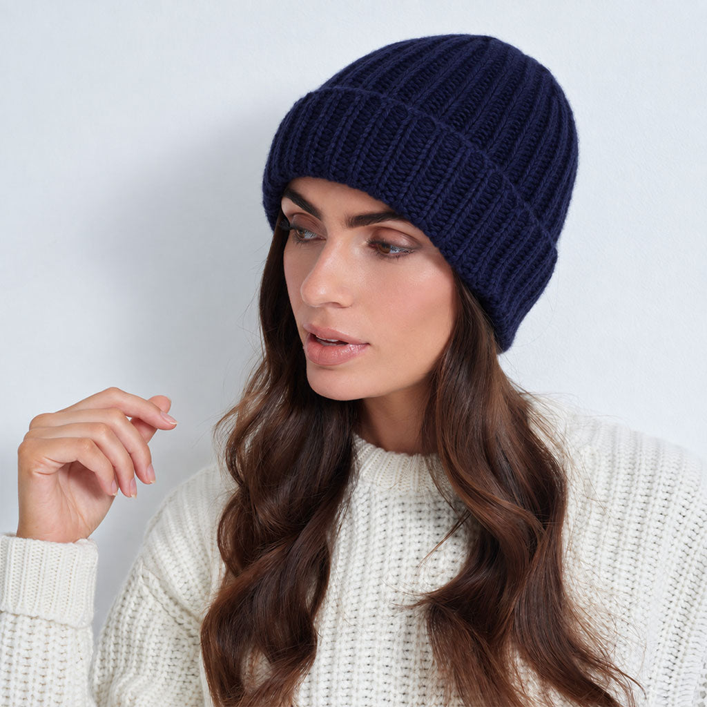 A woman wearing a dark navy blue ribbed knit beanie and a cream-colored knit sweater, shown in a close-up profile shot against a light gray background.