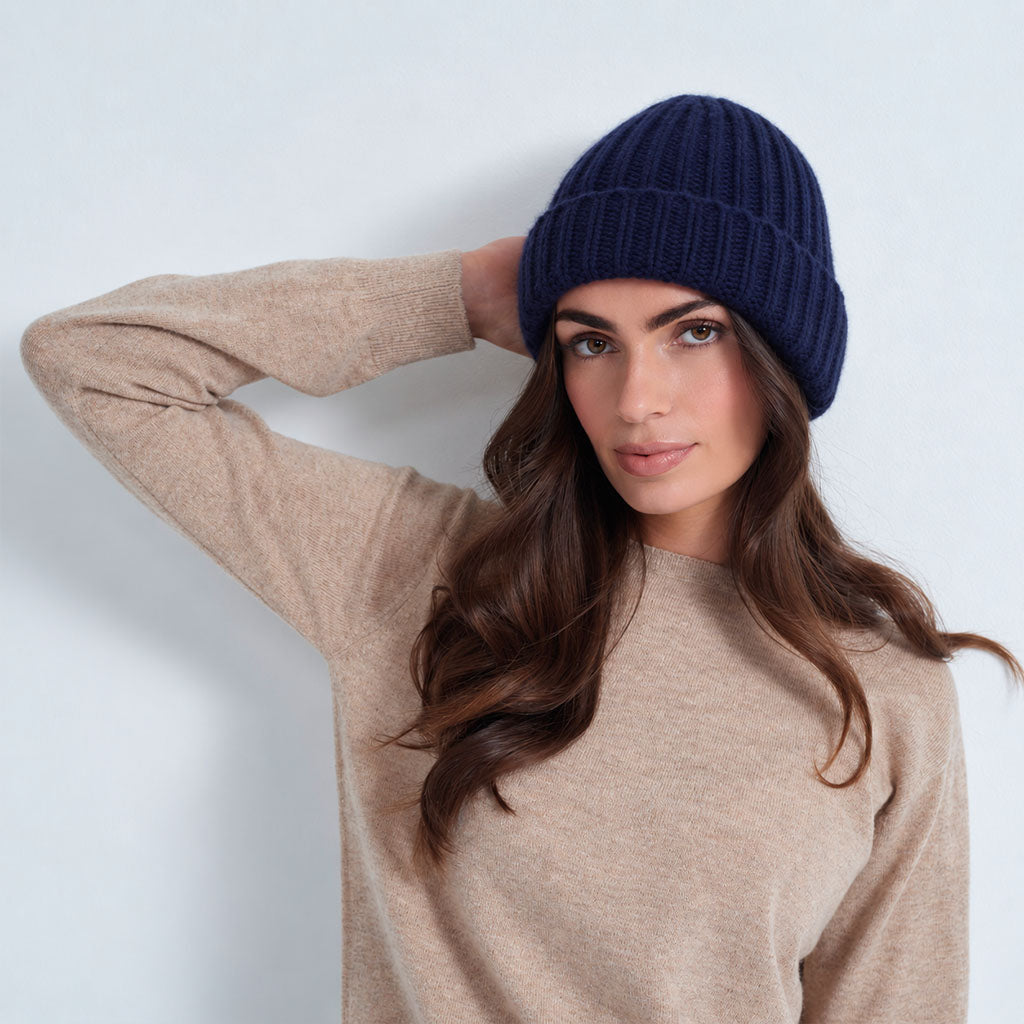 A woman wearing a navy blue ribbed knit beanie and a beige long-sleeve sweater, posing against a light gray studio background.