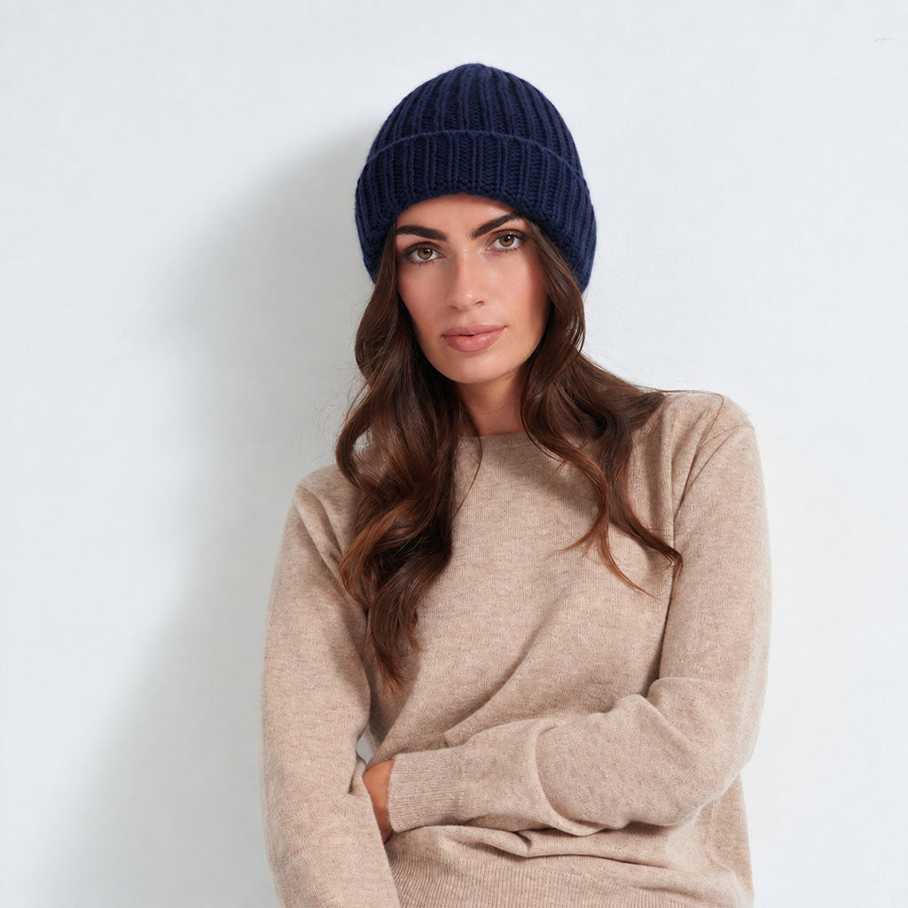 A woman with long wavy brown hair wearing a dark navy blue ribbed knit beanie and a beige knit sweater, posing against a light gray wall.