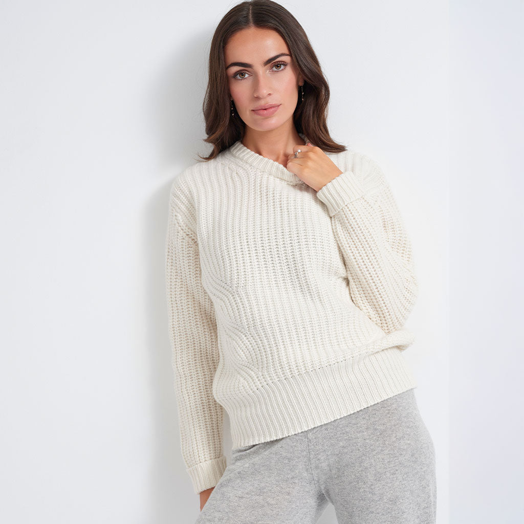 A woman wearing a cream-colored, textured knit crewneck sweater and light gray sweatpants, posing against a plain white wall.