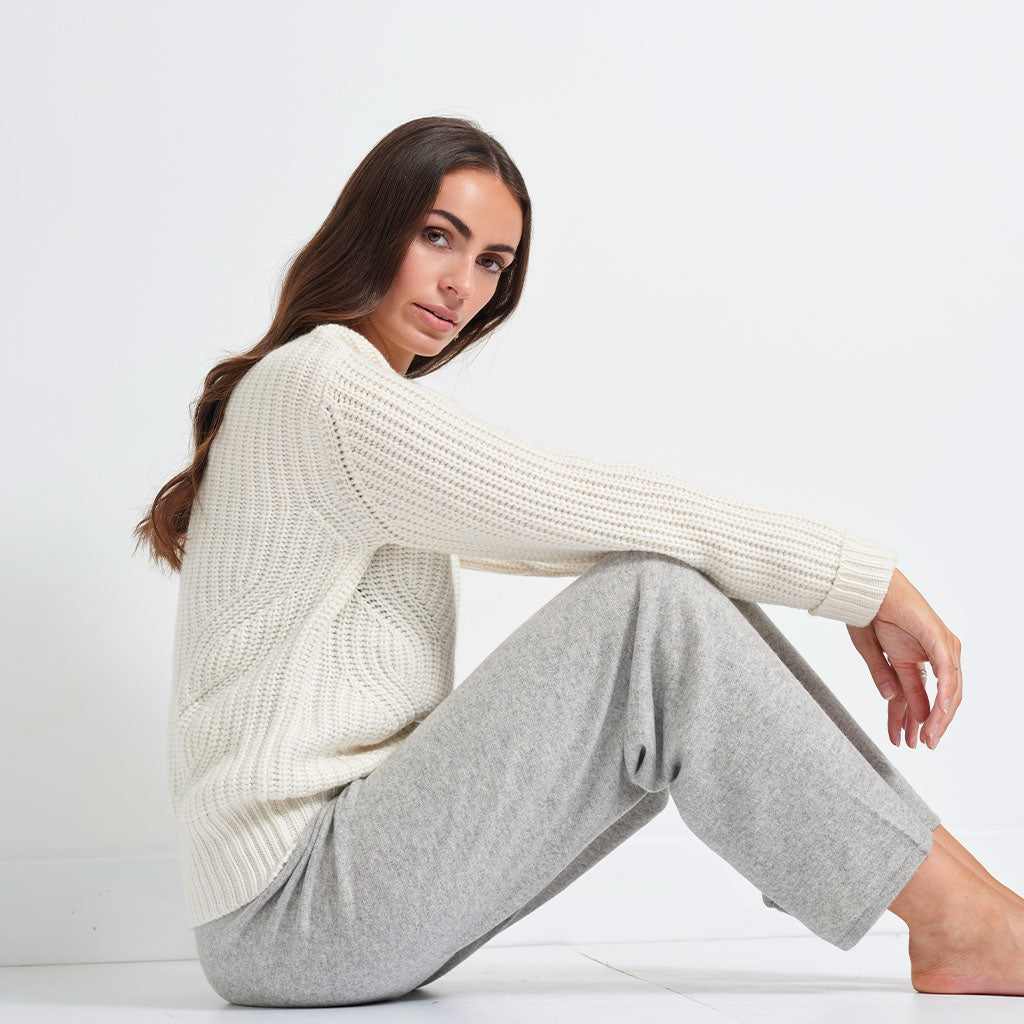 A woman sitting on a white floor against a white wall, wearing a cream-colored textured knit sweater and light gray sweatpants.