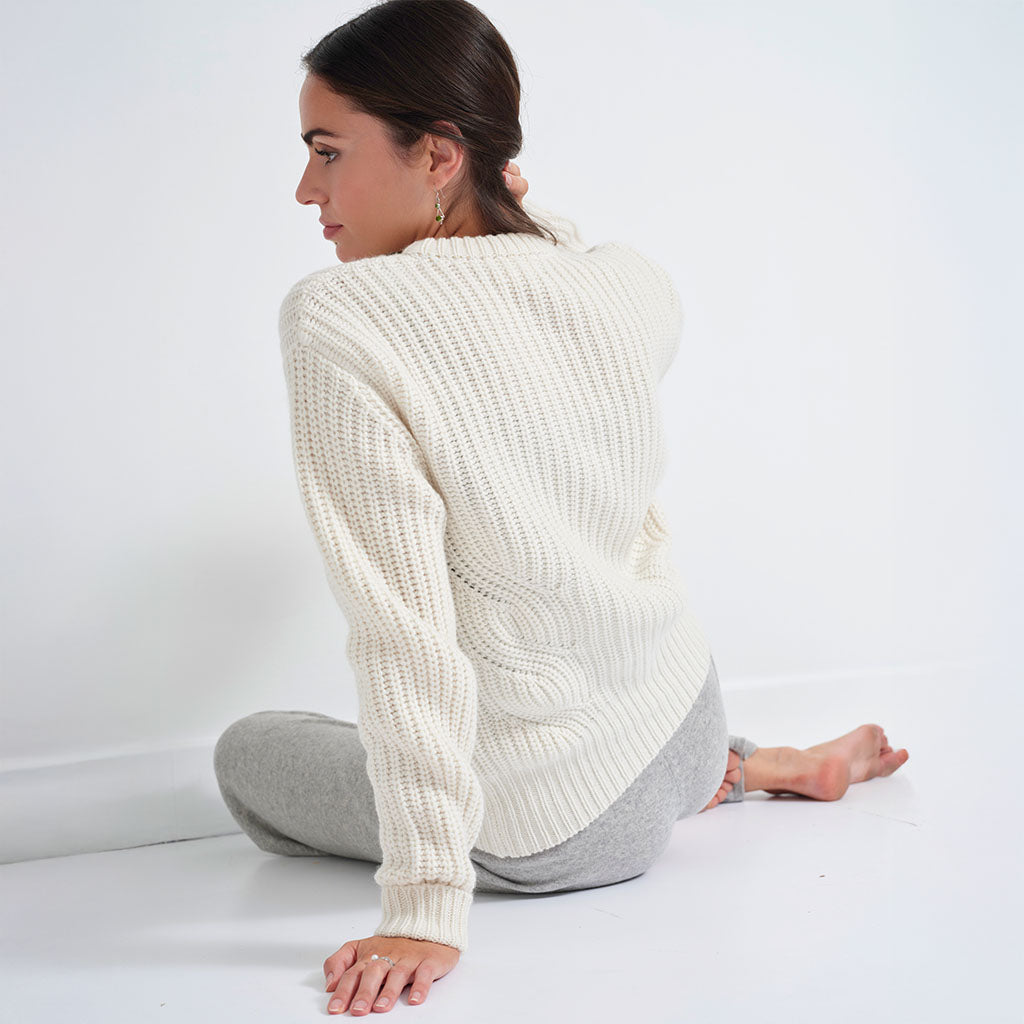 A woman sitting on a white floor against a white wall, wearing a cream cable-knit sweater and gray sweatpants, shown from a back-three-quarter view.