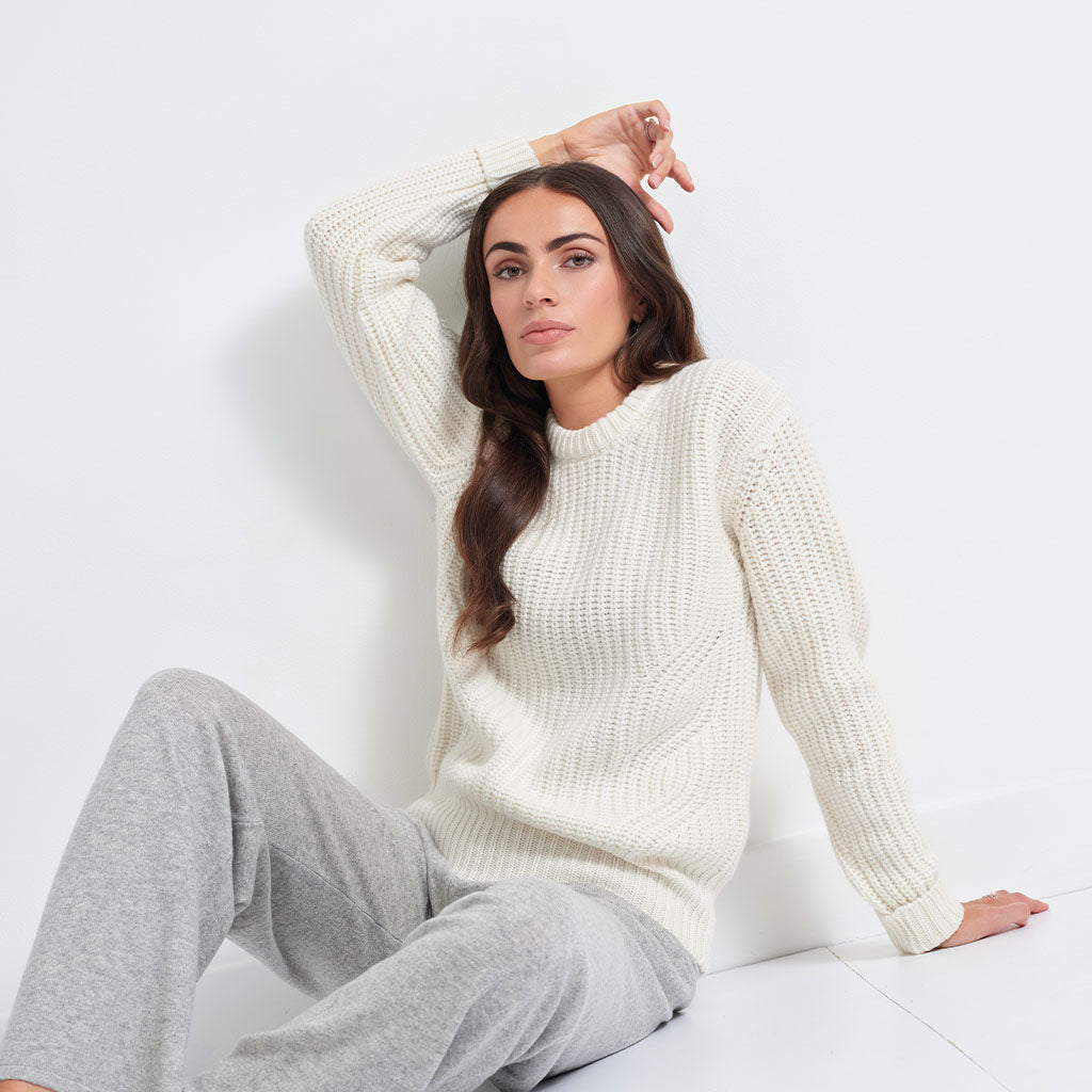 A woman wearing a cream-colored ribbed knit sweater and light gray sweatpants, sitting against a white wall in a studio setting.
