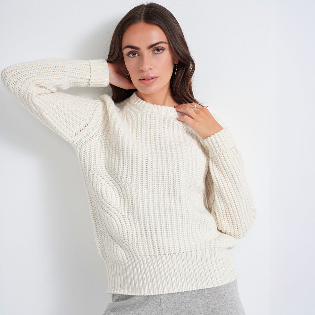 A woman wearing a cream-colored, chunky cable-knit sweater with a crew neck, paired with gray sweatpants, posing against a plain white studio background.
