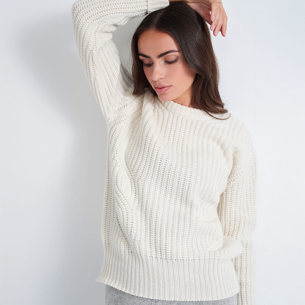 A woman wearing a chunky knit cream-colored sweater with a high neckline and textured cable details, posing against a plain light gray wall.