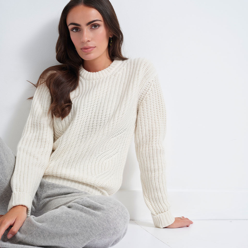 A woman wearing a cream-colored, chunky cable-knit crewneck sweater and gray sweatpants, sitting against a white wall.