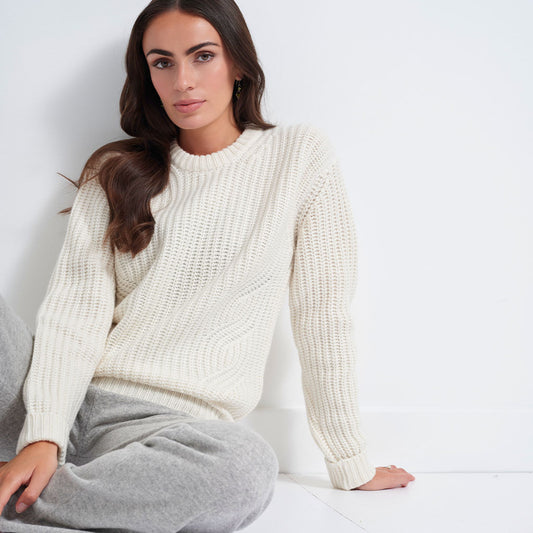 A woman wearing a cream-colored, chunky cable-knit crewneck sweater and gray sweatpants, sitting against a white wall.