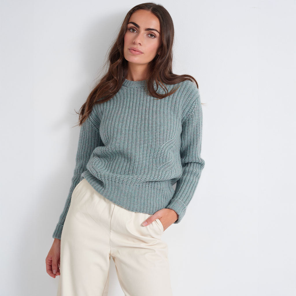 A woman wearing a ribbed knit sweater in a muted sage green color, paired with cream-colored trousers, standing against a plain light gray wall.
