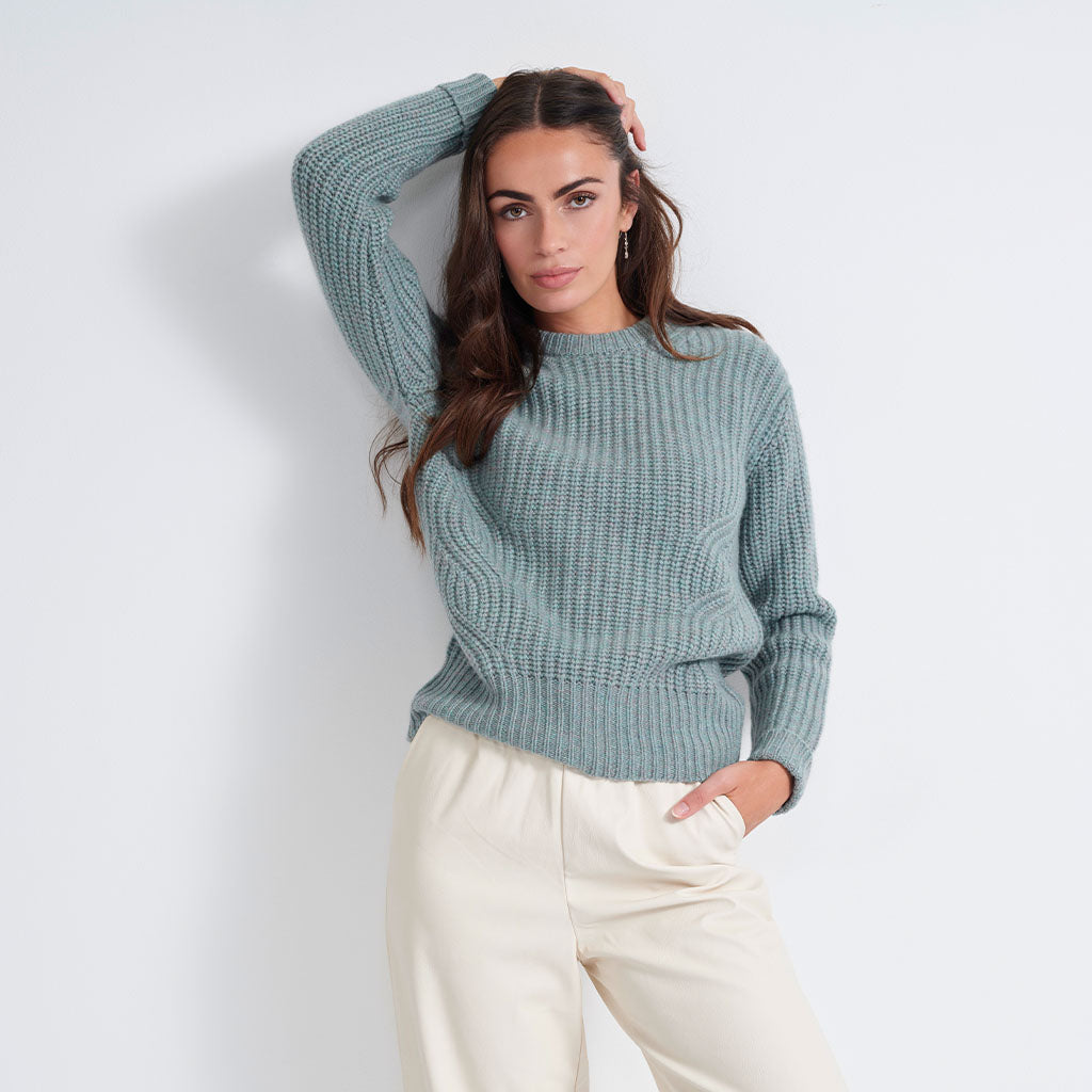 A woman wearing a textured, ribbed knit sweater in a dusty sage green color, paired with cream-colored pants, posing against a light gray studio background.