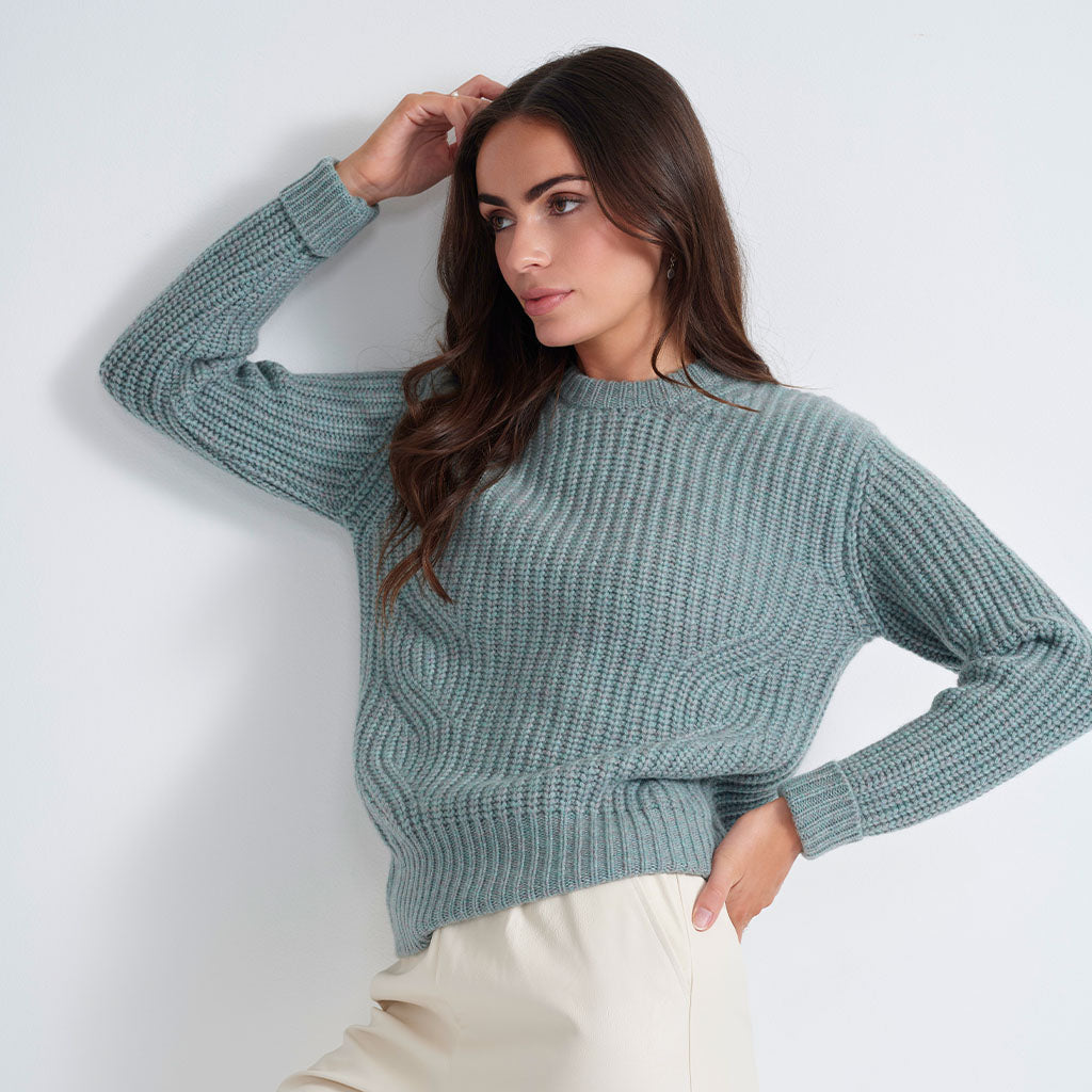 A woman wearing a textured knit crewneck sweater in a muted sage green color, paired with cream-colored trousers, posing against a light gray studio background.
