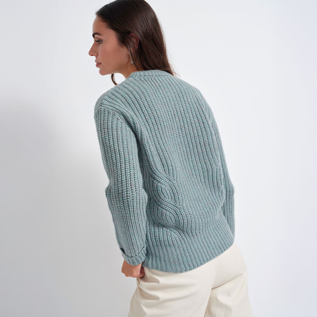 A woman wearing a light blue-green chunky cable-knit sweater and cream trousers, shown from a side-back angle against a plain white studio background.