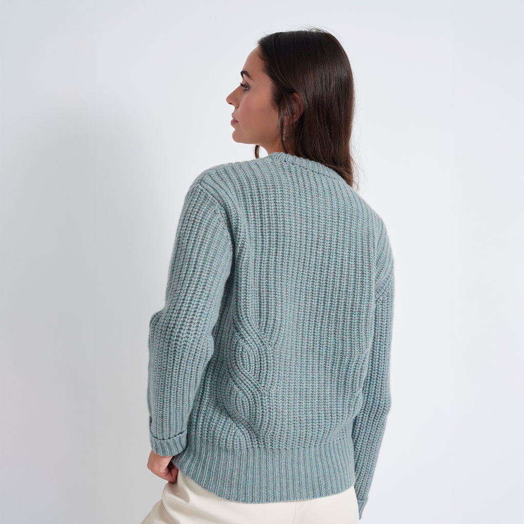 A woman shown from the back wearing a textured, chunky knit cardigan in a dusty sage green color, paired with cream-colored pants, against a plain light gray studio background.