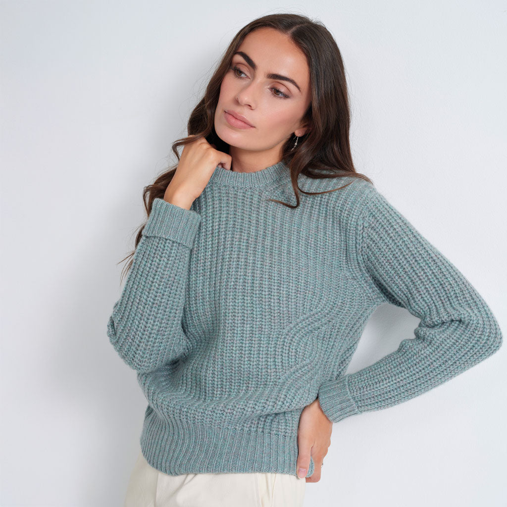 A woman wearing a textured, ribbed knit sweater in a dusty sage green color, paired with off-white trousers, posing against a light gray studio background.