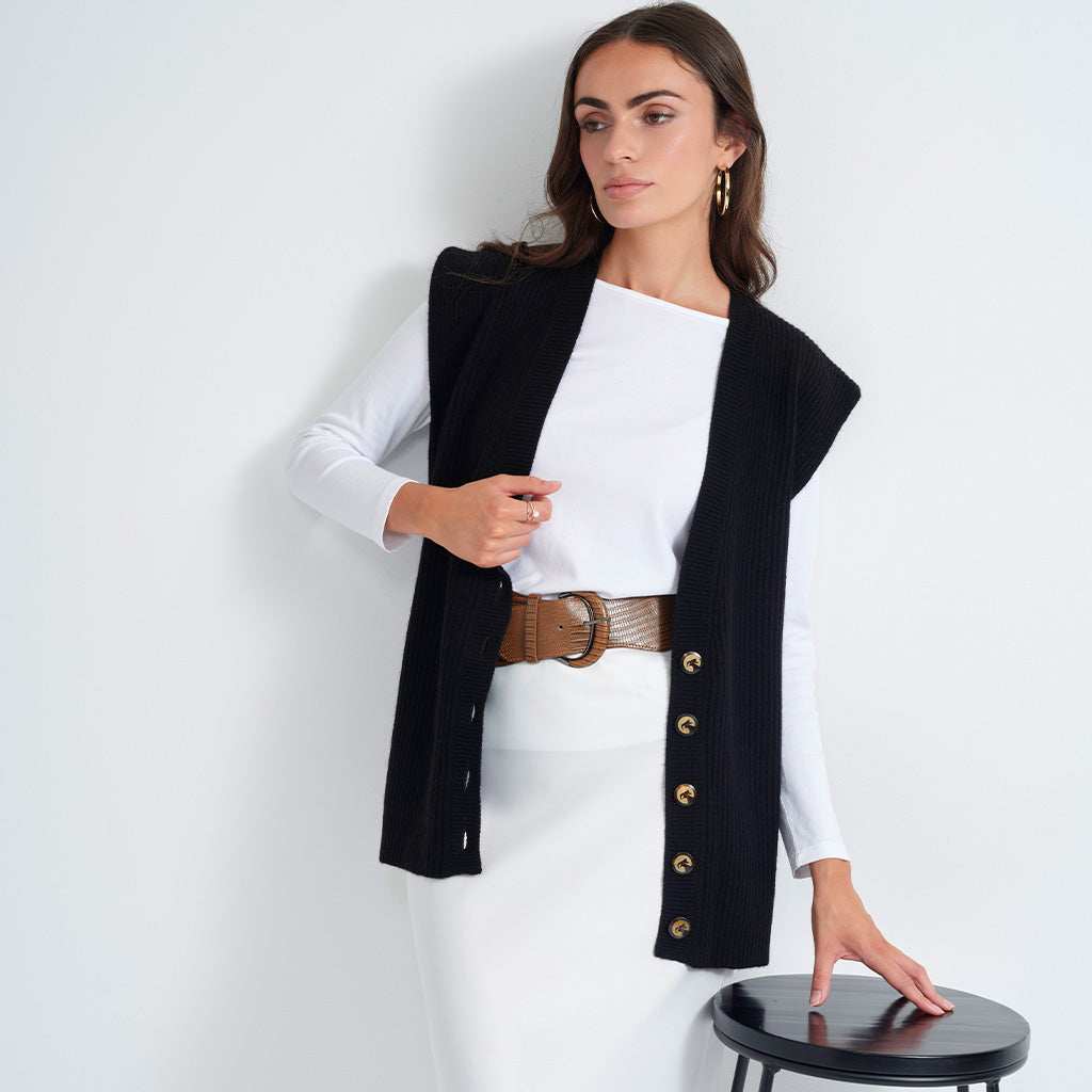 A woman models a black ribbed sleeveless vest with gold-tone buttons over a white long-sleeve top and white skirt, accessorized with a brown belt, standing next to a small black stool against a white wall.