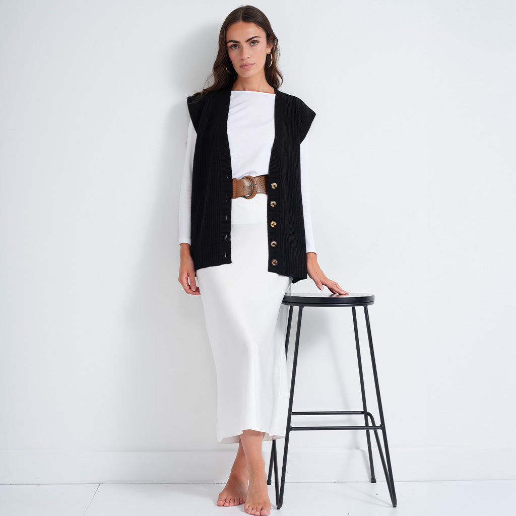 A woman models a black sleeveless vest with gold-tone buttons over a white long-sleeve top and white midi skirt, accessorized with a brown belt, standing next to a black metal stool against a white wall.