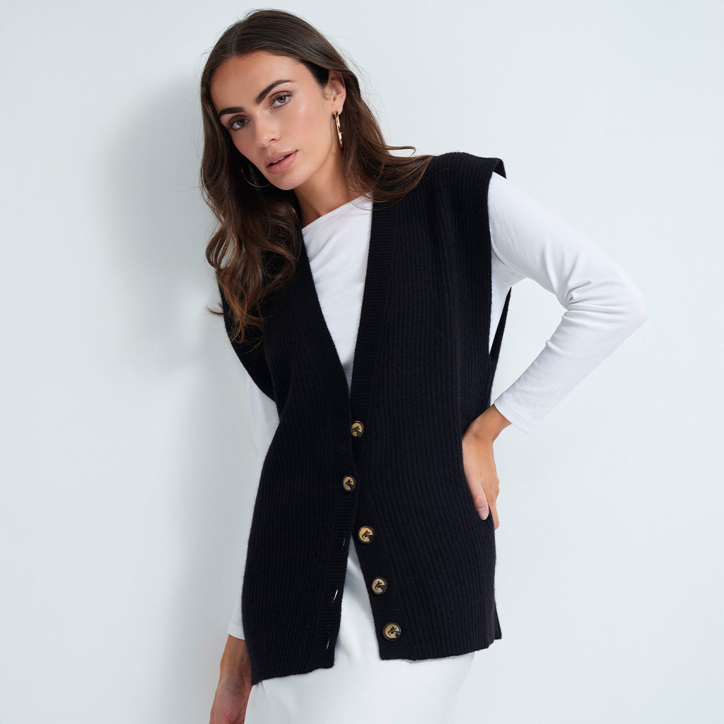 A woman wearing a black ribbed knit vest with gold buttons over a white long-sleeve top, posing against a light gray studio background.