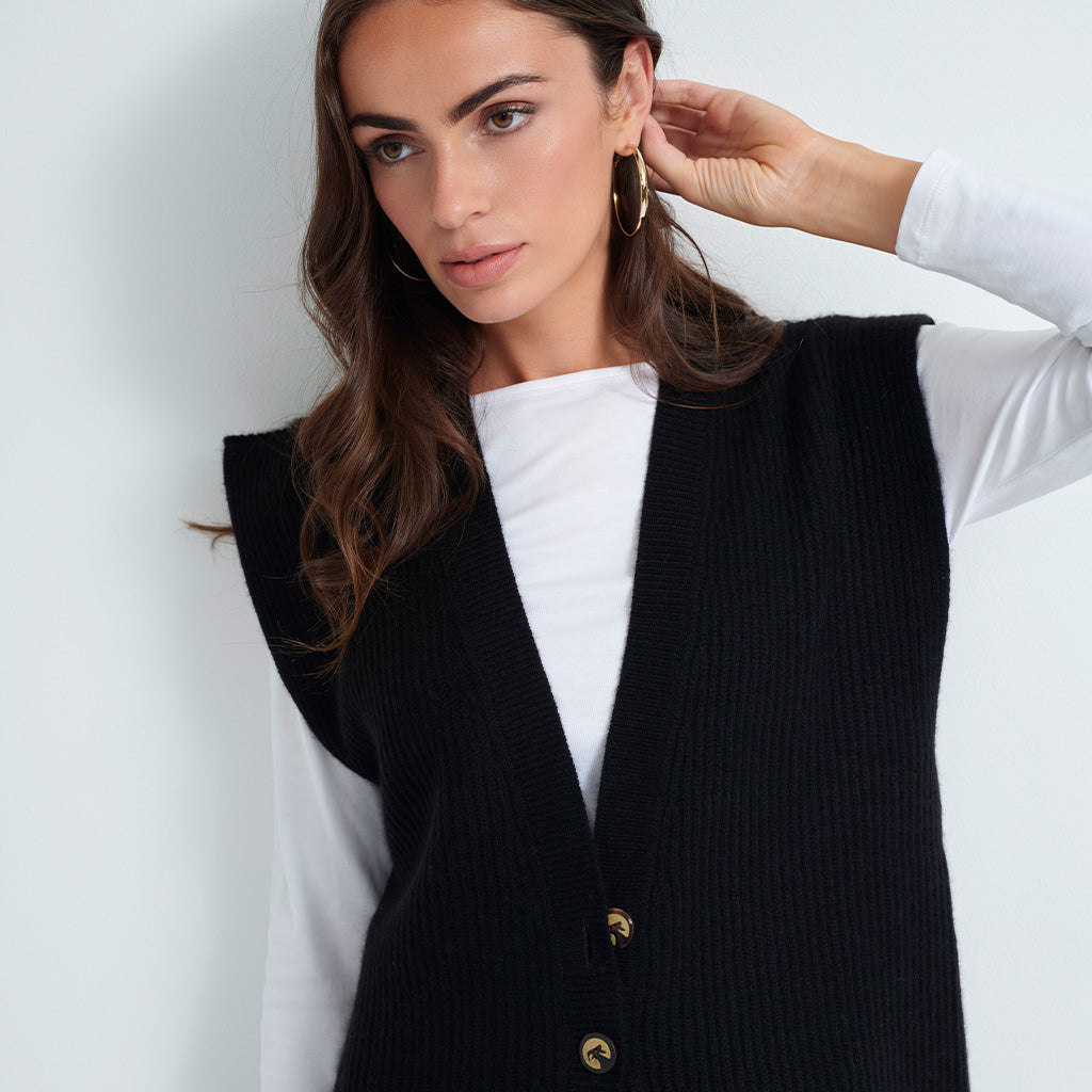 A woman wearing a black ribbed knit vest over a white long-sleeve top, with gold hoop earrings, photographed against a light gray studio background.
