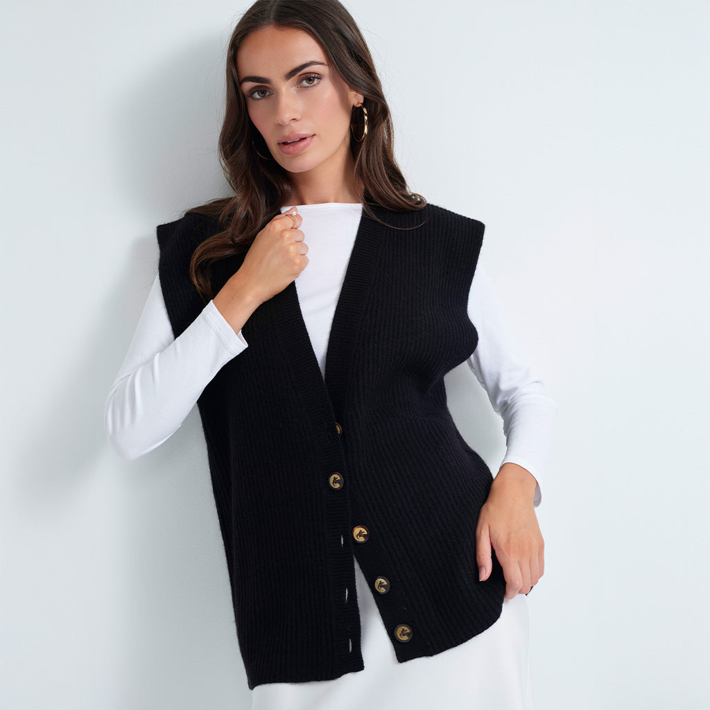 A woman wearing a black ribbed knit vest over a white long-sleeve top, standing against a light gray studio background.