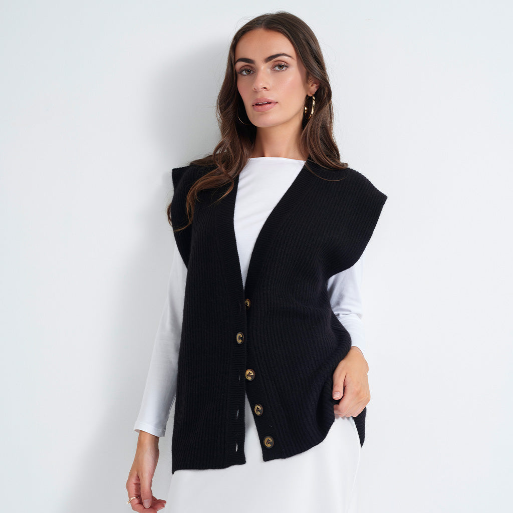 A woman wearing a black ribbed knit sleeveless vest with gold buttons over a white long-sleeve top, photographed against a plain light gray studio background.
