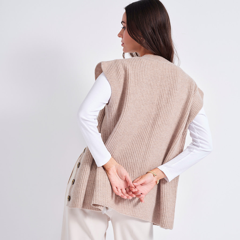 A woman shown from the back wearing a sleeveless beige ribbed knit vest over a white long-sleeve top and light pants, against a plain light background.