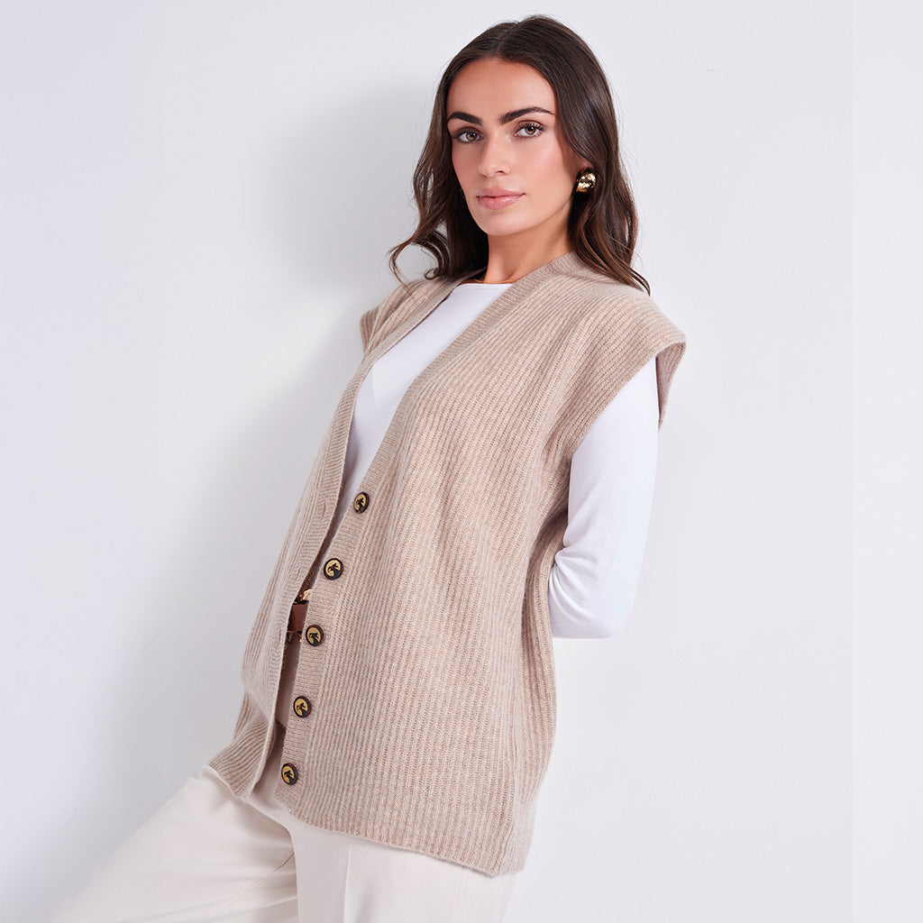 A woman models a sleeveless beige ribbed knit vest with brown buttons over a white long-sleeve top and white pants, set against a plain light gray studio background.