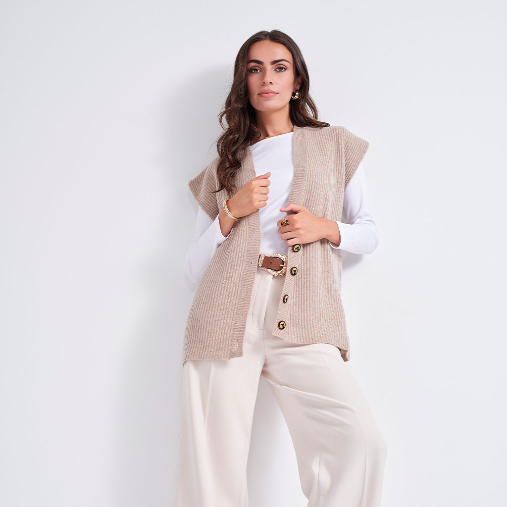 A woman models a sleeveless beige ribbed knit vest with gold-tone buttons over a white long-sleeve top and cream wide-leg trousers, set against a plain white studio background.