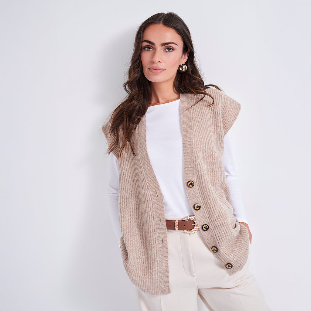 A woman modeling a sleeveless beige ribbed knit vest with gold-tone buttons over a white long-sleeve top and cream pants, against a plain light gray studio background.