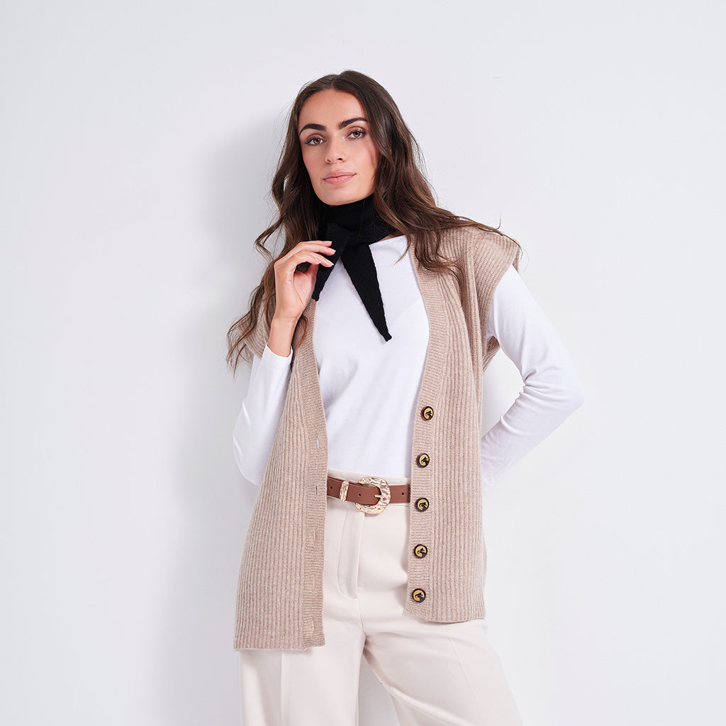 A woman modeling a sleeveless beige ribbed knit vest with gold buttons over a white long-sleeve top and cream pants, against a plain light gray studio background.