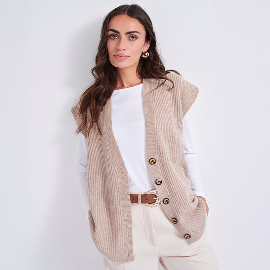 A woman models a sleeveless beige ribbed knit vest with gold-tone buttons over a white long-sleeve top and cream pants, set against a plain light gray studio background.